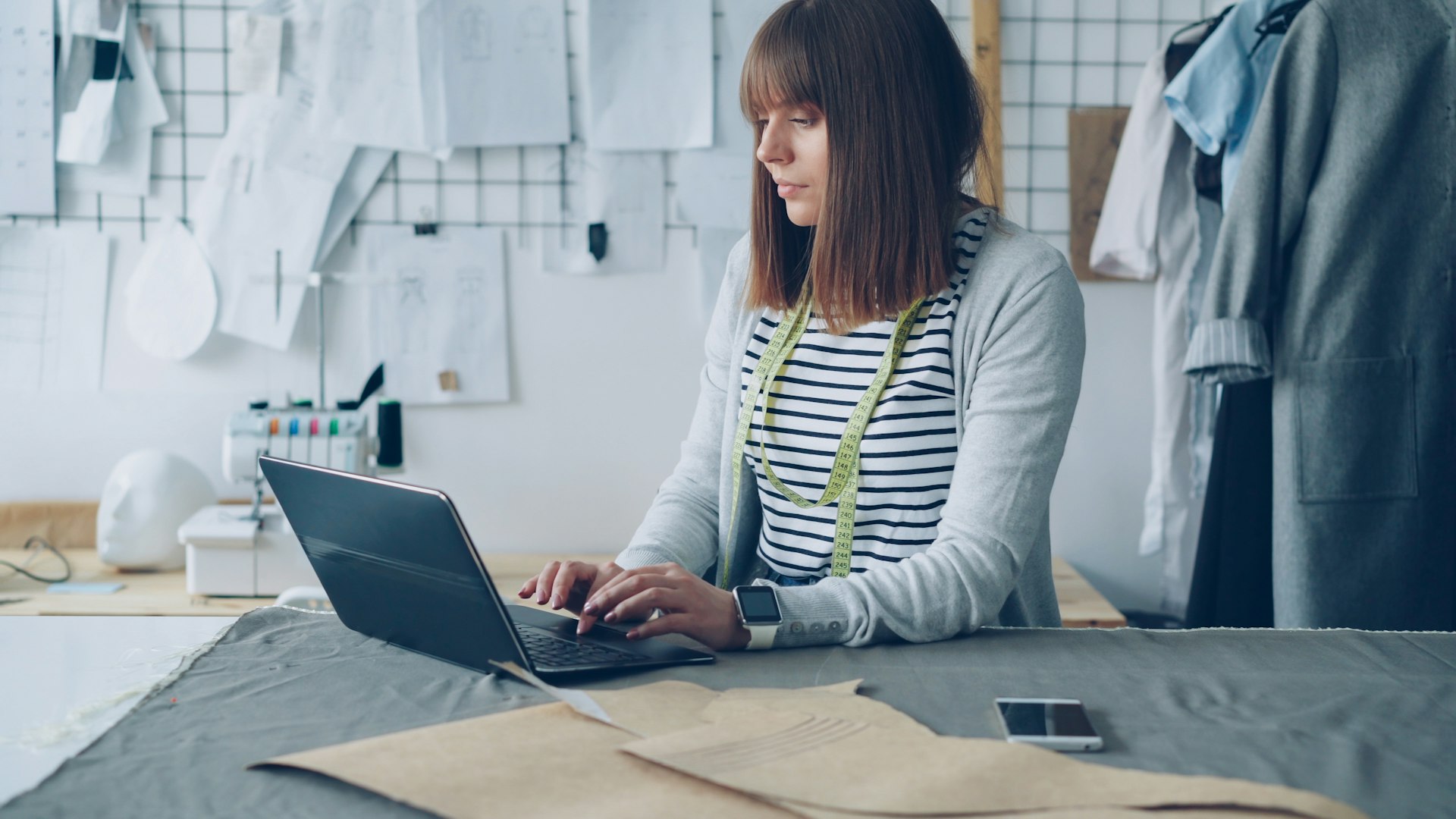 A seamstress works on her laptop in a bright workspace.